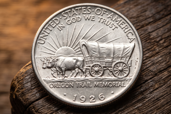 Close-up of a 1926 Oregon Trail Memorial Half Dollar showing oxen pulling a covered wagon, with a rising sun and 'UNITED STATES OF AMERICA' and 'IN GOD WE TRUST' above, all on a rustic wooden surface.